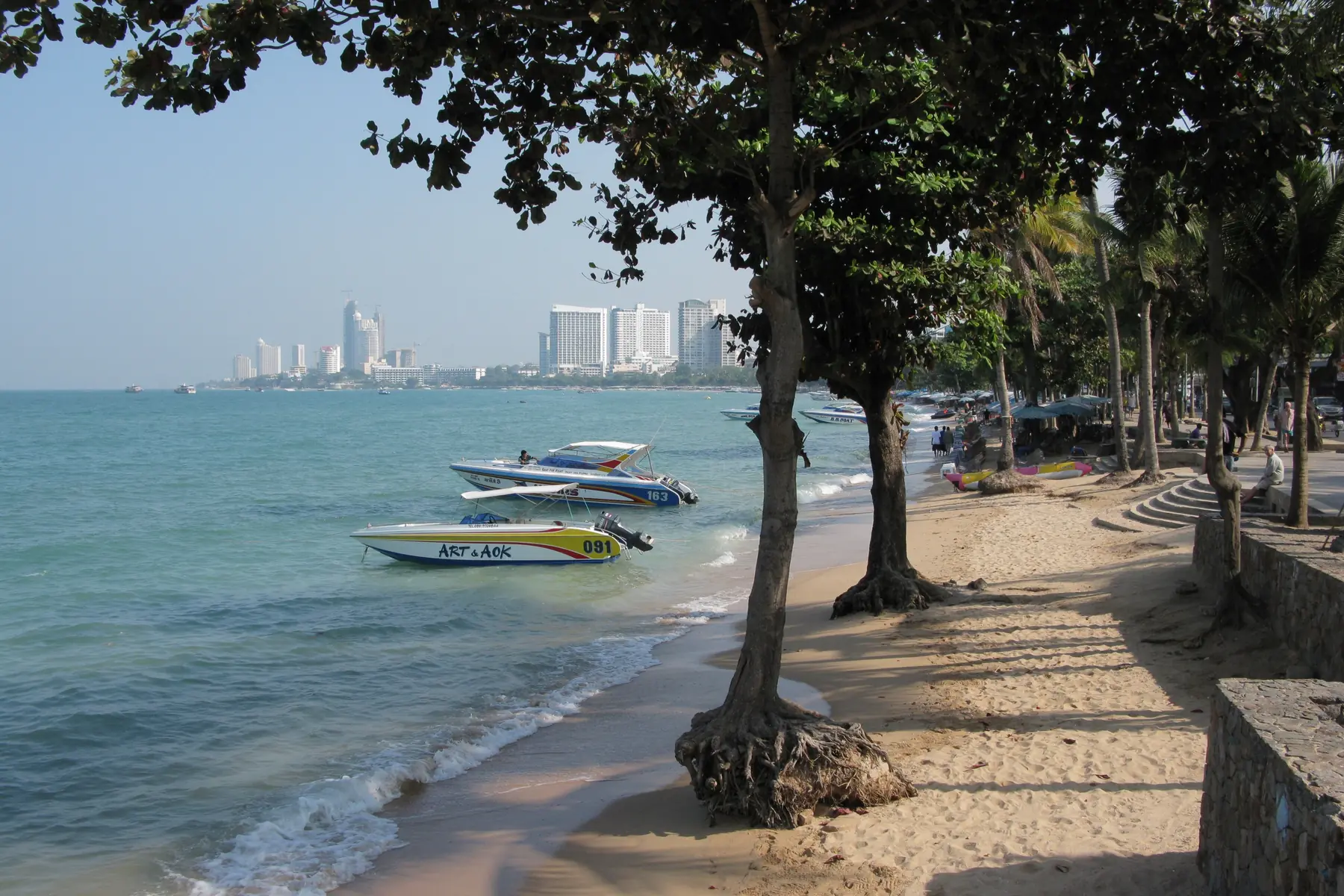 Træer langs stranden i Pattaya med speedbåde i det turkise vand og hoteltårne i det fjerne